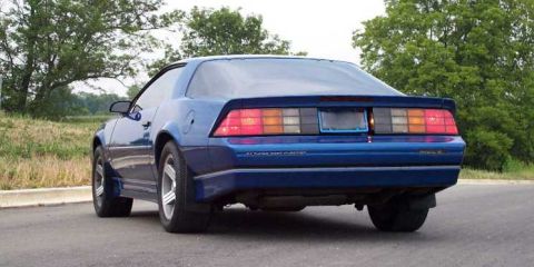 1998 Ford Crown Victoria - Rear view of a blue coupe, possibly a Chevrolet Camaro, parked on a roadside with trees in the background.