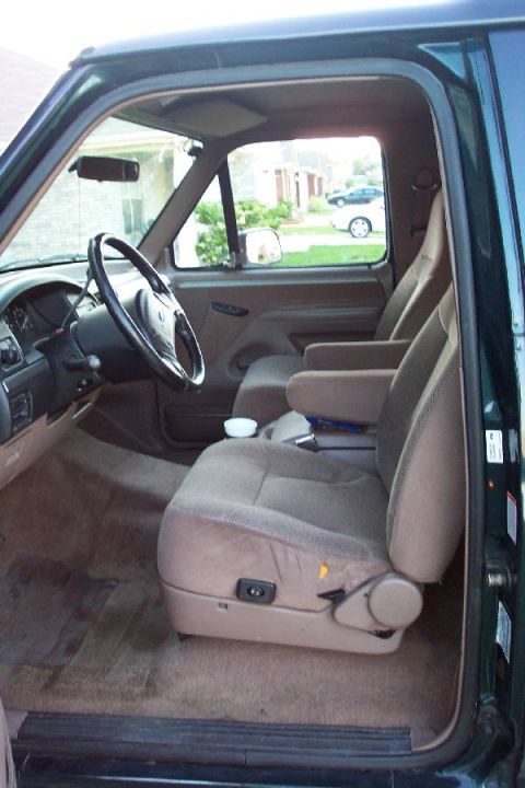 1992 Ford Bronco - Interior of a green Ford Bronco viewed through the open driver's door, showing tan cloth captain's-style front buckets, a center console, m…
