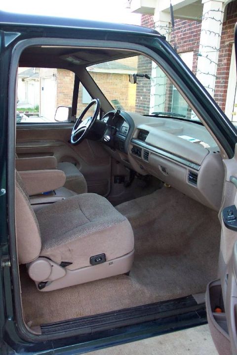 1992 Ford Bronco - Interior of a green SUV shot through the open passenger door, showing the tan dash with center vents, a four-spoke steering wheel, and tan…