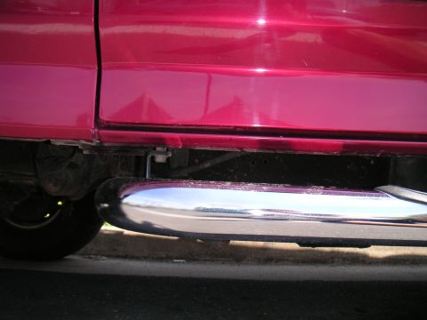 1994 Ford Bronco - Low-angle close-up of a chrome tube side step running beneath the red rocker panel and door of a truck, with dust collected on the polished…