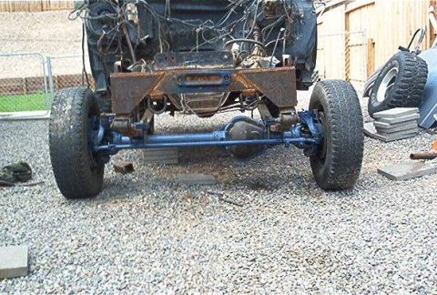 1986 Ford Bronco - Front view of a bare truck chassis with a freshly painted blue front axle and leaf springs, mounted on chunky off-road tires. A rusty frame…