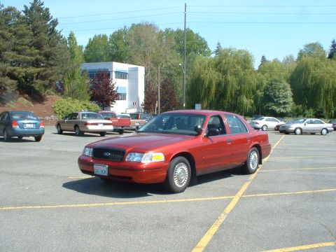 1999 Ford Crown Victoria - A red sedan is parked in a lot with other vehicles and a building in the background.