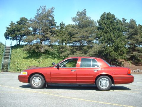 1999 Ford Crown Victoria - A red 1999 Ford Crown Victoria is parked on asphalt with trees and a grassy hill in the background.
