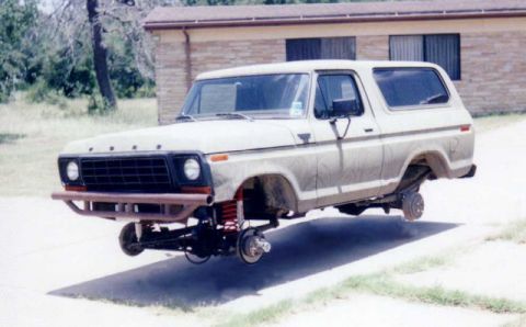 1990 Ford Bronco - A white late-1970s full-size Ford Bronco sits on jack stands with all four wheels removed, showing the front axle, red coil springs and bra…