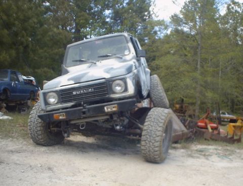 1989 Ford Bronco - A Suzuki Samurai is shown with a camouflage paint job and significantly modified suspension, parked in an outdoor lot with other vehicles.