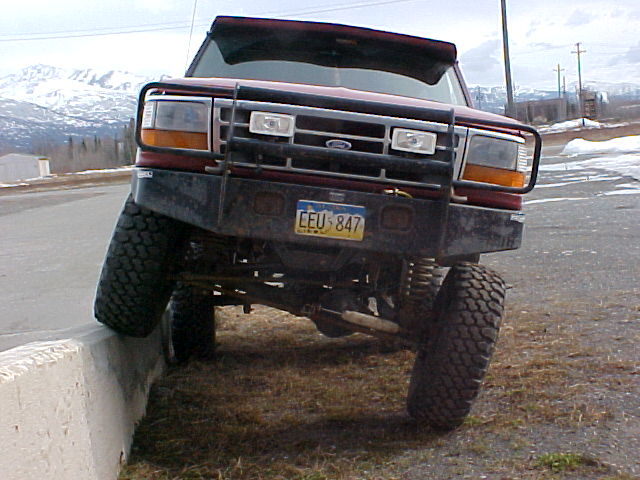 Lifted red Ford pickup front end perched on a concrete curb with snowy mountains behind