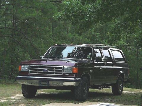 1988 Ford C-350 (Centurion Conversions) - A Ford Bronco sits parked on a dirt path surrounded by trees.