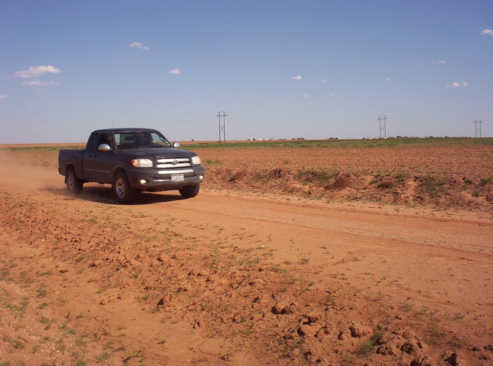 Dark gray Toyota Tundra driving on a dirt road, kicking up dust. Open field and power lines in background