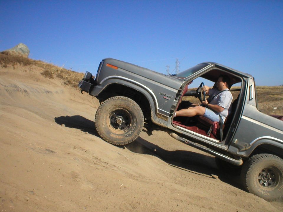 Gray topless Ford Bronco climbing a dirt hill with front wheel lifting, driver inside