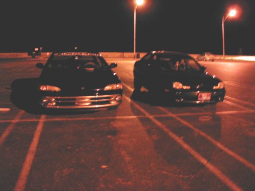 Two dark sedans parked at night, one in the foreground with a chrome grille