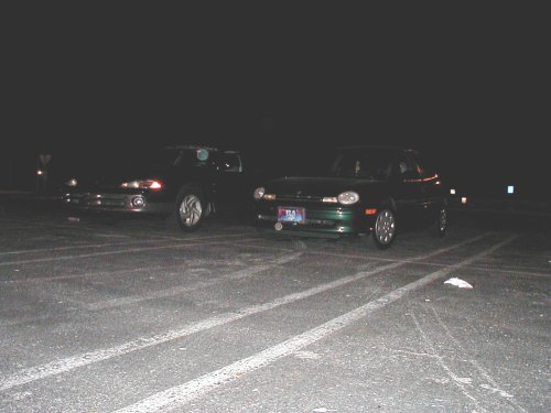 Two cars parked in a lot at night. The car on the right is dark green