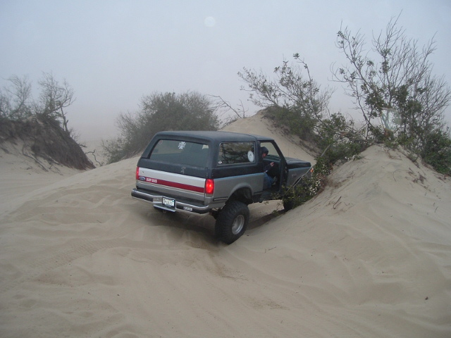 Two-tone black and silver full-size Ford Bronco parked in foggy sand dunes, rear three-quarter view