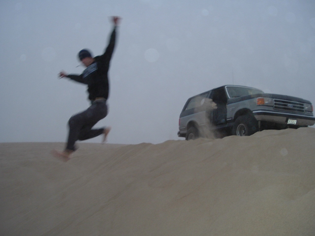Person leaping off a sand dune beside a gray Ford Bronco at dusk