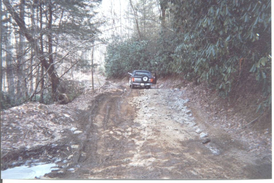 1984 Ford Bronco driving on a muddy, rocky off-road trail through a forest