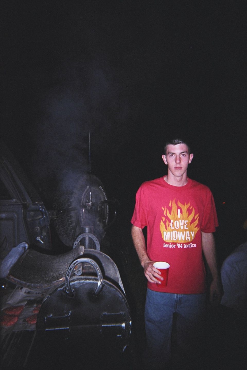 Man in red t-shirt holding red cup, standing by barbecue grill with smoke. Text on shirt reads 'I LOVE MIDWAY Senior '84 Bonfire'