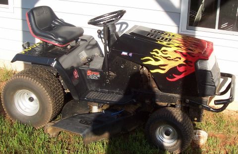 1979 Ford Bronco - A black riding lawnmower with flame graphics is parked on grass next to a house.