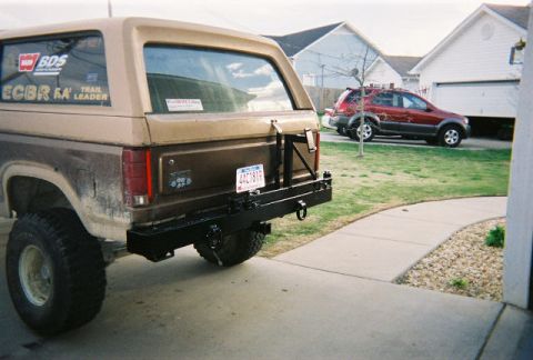 1983 Ford Bronco - The rear of a classic SUV is shown with a custom bumper and spare tire mount, while another vehicle is parked in the background.