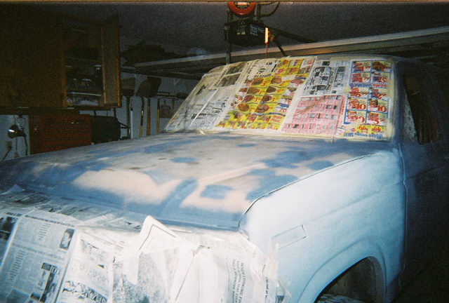 Truck hood and fender in gray primer with windshield masked in newspaper during home bodywork