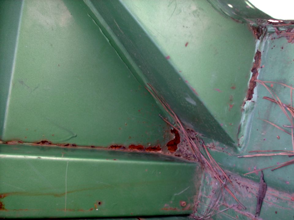 Close-up of a green vehicle panel with significant rust damage along the seam and in a corner. Pine needles are visible in the rusted area