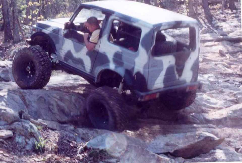 Camouflaged Suzuki Samurai off-roading over rocks, driver visible
