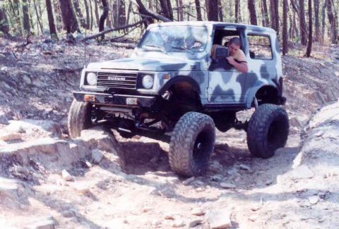 1989 Ford Bronco - A Suzuki Samurai with camouflage paint is shown off-roading on a dirt path with large rocks.