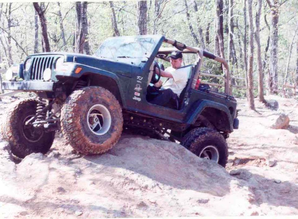 Green Jeep Wrangler climbing a dirt hill with large tires. Driver is visible