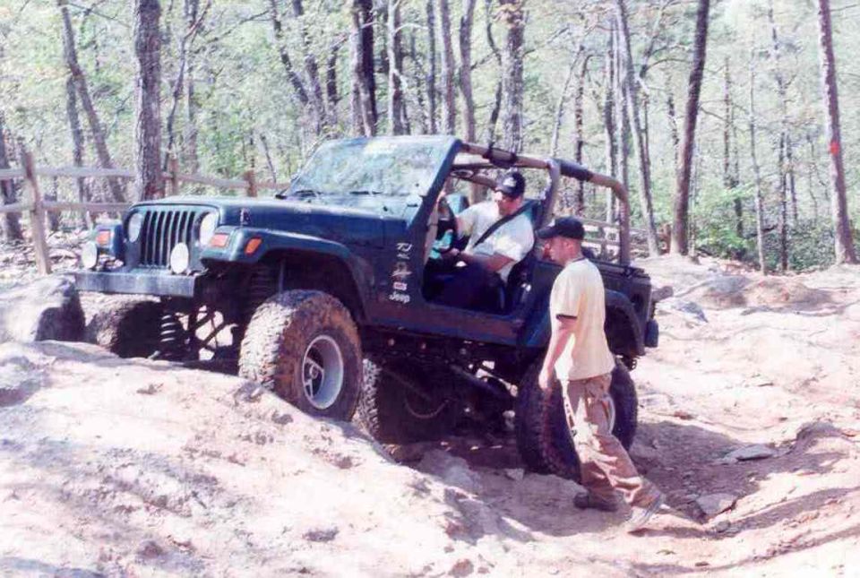 Dark green Jeep Wrangler off-roading on a dirt trail, with two men standing nearby