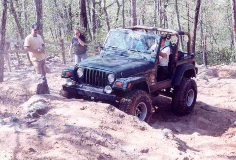 1989 Ford Bronco - A dark green Jeep Wrangler TJ is shown off-road on a rocky trail with two people standing nearby.
