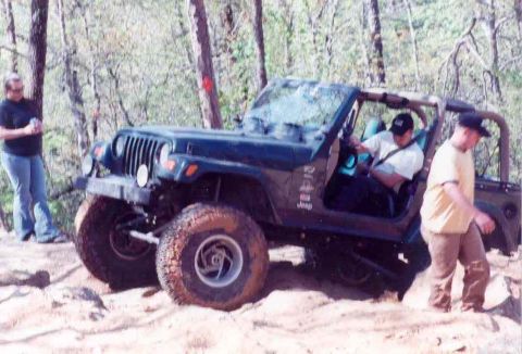 1989 Ford Bronco - Jeep Wrangler navigating a rough, wooded terrain with two individuals observing.