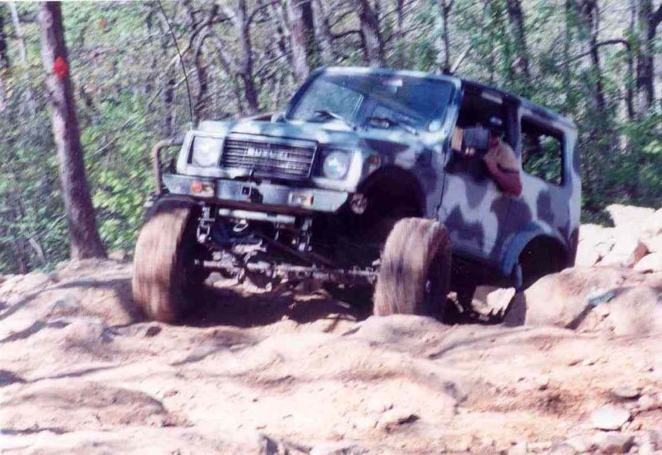 Camouflaged Suzuki Samurai off-roading on rocky terrain, front-left tire lifted