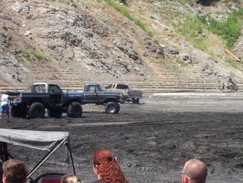 2000 Ford F-450 - Two lifted blue-and-gray Ford pickups on oversized mud tires sit side by side on a wet, churned mud pit at an off-road event, one with a ro…