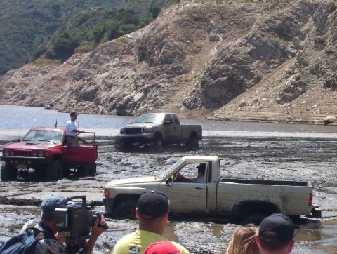 2000 Ford F-450 - Several trucks are stuck in the mud near a lake, with a rocky hill and trees in the background.