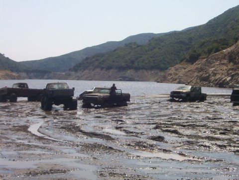 2000 Ford F-450 - Multiple trucks are stuck in a muddy area next to a large body of water, with hills in the background.