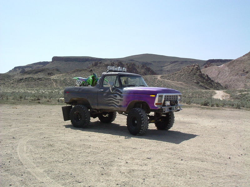 Lifted black and purple flamed 4x4 truck on dirt with dirt bike in bed, desert hills behind