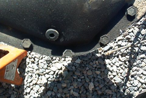 1986 Ford Bronco - Close-up of a differential housing with visible bolts, resting on gravel with an orange jack nearby.