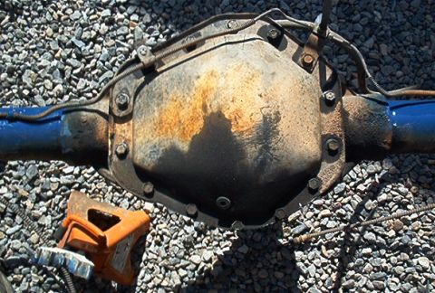 1986 Ford Bronco - A rear axle differential assembly is shown with blue axle tubes and an orange jack stand resting on gravel.