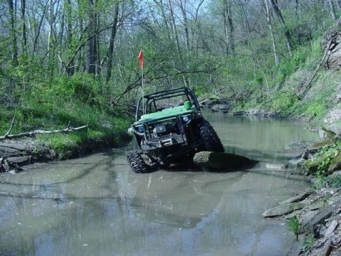 1992 Ford Bronco - An off-road vehicle navigates through a muddy stream surrounded by trees, with an orange flag visible on its antenna.