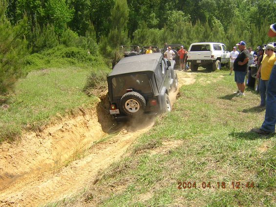 Silver Jeep Wrangler climbing out of a dirt trench at an off-road park with spectators watching
