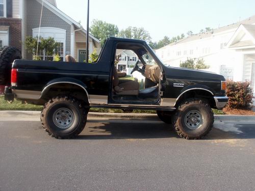 Black lifted Ford Bronco SUV, side view, big mud tires, door open, parked on street