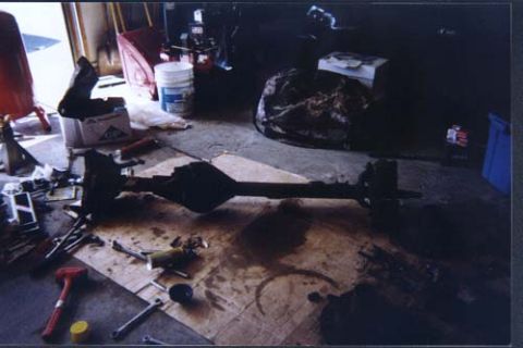 1996 Ford Bronco - A bare solid rear axle housing sits on a cardboard-covered garage floor with an oil stain spreading beneath it, its differential center sec…