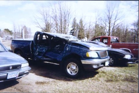 1996 Ford Bronco - This severely damaged blue pickup truck is parked outside with other vehicles under a cloudy sky.