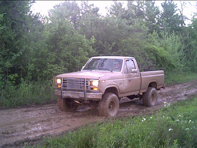 Lifted mud-covered Ford F-150 pickup with headlights on driving through a muddy wooded trail