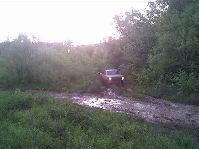 Pickup truck with lights on emerging from brush onto a muddy trail in dim evening light