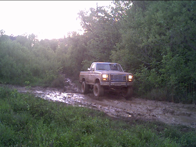 Lifted tan Ford pickup with brush guard on a muddy trail, headlights on at dusk