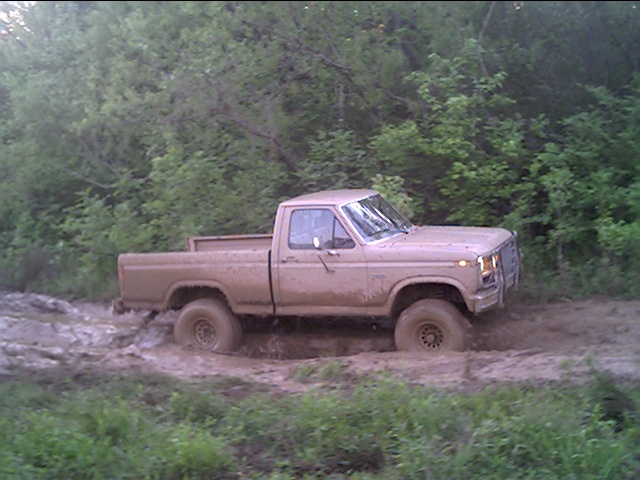 Mud-covered tan Ford pickup, side profile, parked in a muddy trail in the woods