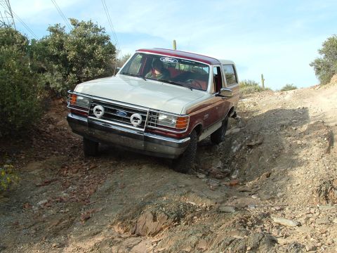 1990 Ford Bronco - A two-tone Ford Bronco navigates a challenging off-road path under a bright sky.