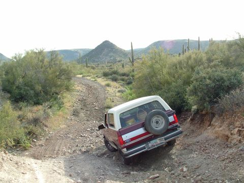 1990 Ford Bronco - A Ford Bronco navigates a rough, rocky desert trail with saguaro cacti and mountains in the background.