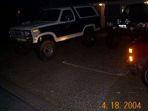 1986 Ford Bronco - A two-tone Ford Bronco is parked on a gravel surface at night, with a bent metal bar lying in front of it.