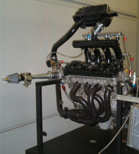 1985 Ford LTD Crown Victoria - A turbocharged V8 engine mounted on a black tubular display stand against a white wall, with polished stainless headers feeding a turbochar…