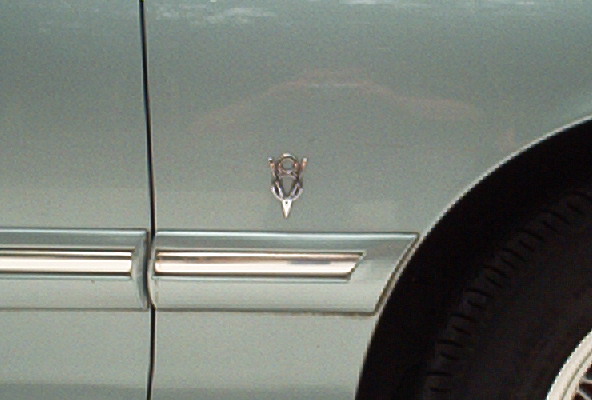 Light blue car fender with chrome trim and a V8 emblem. Tire visible on the right